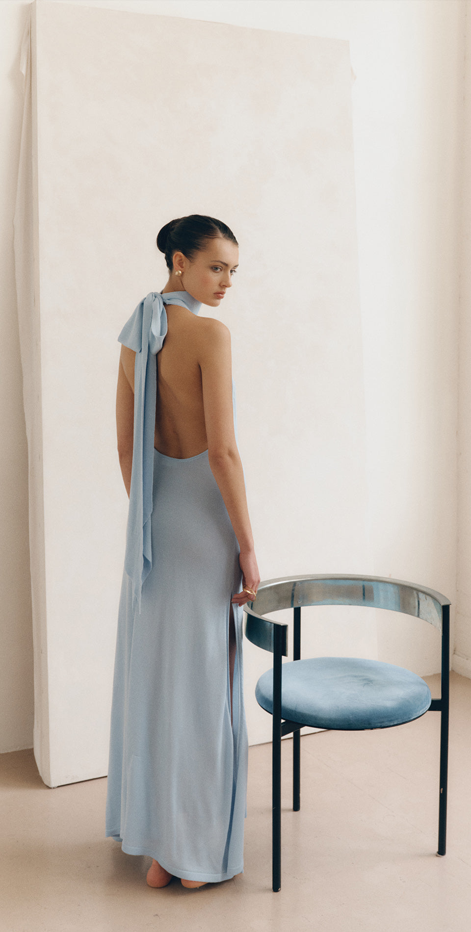 Model wearing the LENY Cascade Dress 2.0 in Aquamarine, a blue backless maxi dress with halter tie detail.