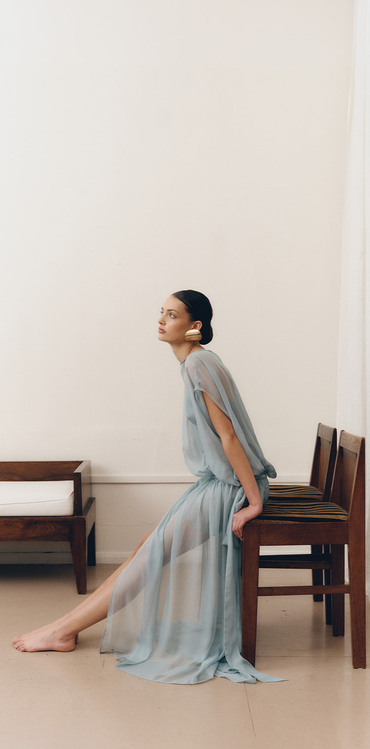 Model seated in the LENY Celeste Sheer Maxi Dress in Storm Blue, showing the soft sheer fabric and relaxed fluid silhouette.